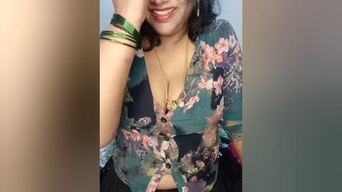 Media: A video of a smiling woman with medium skin tone, wearing a floral-patterned blouse, gold bangles, and a necklace, posing indoors against a plain background.