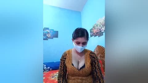 Video of a woman with light skin, dark hair, wearing a white face mask, yellow cardigan, black bra, and floral-patterned blanket in a blue-walled room with framed pictures.