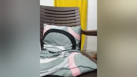 Media: Video of a brown wicker chair with a geometric-patterned green, pink, and black cushion, placed against a white wall with a yellow curtain.