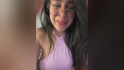 Video of a young woman with long black hair, wearing large round glasses and a light pink halter top, smiling warmly. Background is blurred, focusing attention on her face.