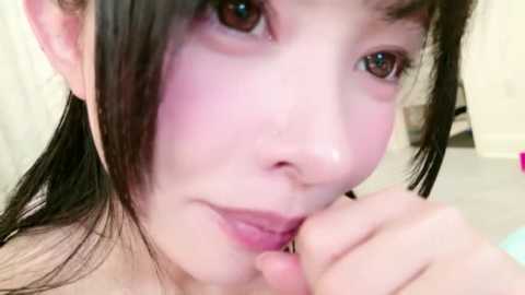 Video of a young East Asian woman with pale skin and straight black hair, lightly blushing. She gazes into the camera with a gentle expression, her lips slightly parted. The background is blurred, featuring a light-colored indoor setting.