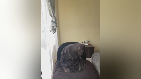 Video of a messy bedroom with a brown comforter and pillows, beige walls, a white door, a dark wooden nightstand with toiletries, and a small window with light green curtains.