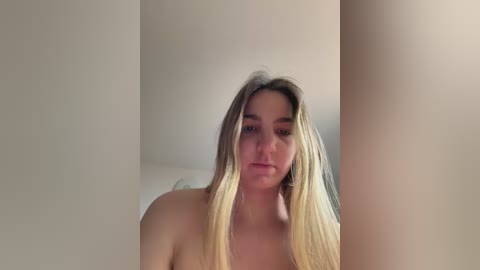 A video of a nude, light-skinned woman with long, straight blonde hair, looking directly at the camera with a neutral expression. The background is plain and off-white, creating a minimalist setting.