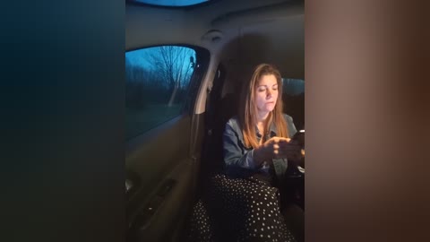 Video of a young woman with long blonde hair, wearing a denim jacket and black skirt, seated in a dimly lit car at night, texting on her phone.
