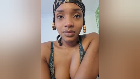 Media: Video of a young Black woman with medium brown skin, wearing a leopard-print headscarf, green bra, and hoop earrings, smiling slightly. She is standing in front of a white wall and green curtain.