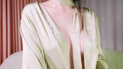 Video of a woman with pale skin and dark hair wearing a light green, V-neck blouse, partially unbuttoned, revealing cleavage. Background features soft, vertical pink and beige curtains.