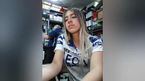 Video of a young woman with long, straight blonde hair, wearing a white and blue soccer jersey with \"FOX\" text. She sits in a cluttered room with shelves and boxes in the background.