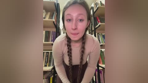 Video of a young woman with long brown hair in braids, wearing a beige top and brown overalls, leaning forward, surrounded by bookshelves filled with colorful books.