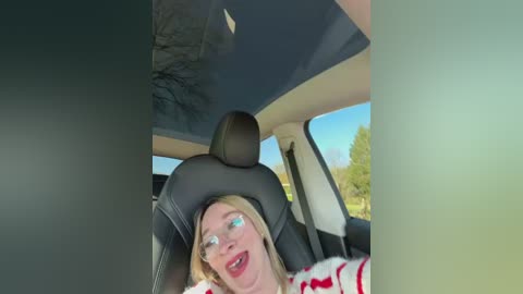Video of a blonde woman in glasses, wearing a white and red striped shirt, sitting in a car with a black headrest, looking out the window.