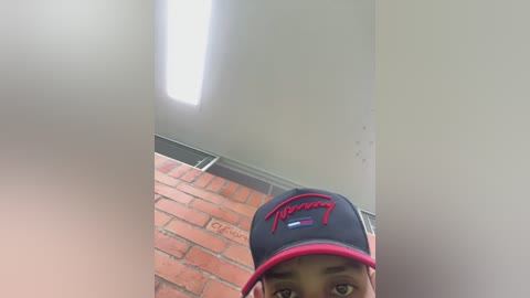 Video of a young Black man in a black cap with red stitching, standing in a brick-walled room with a ceiling light.