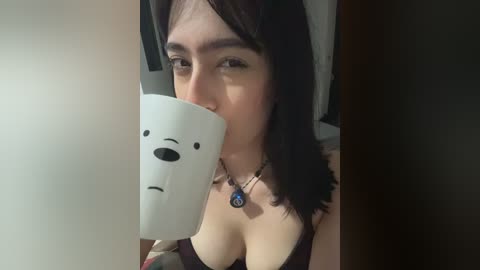Video of a young woman with fair skin, dark hair, and a necklace, drinking from a white cup shaped like a sad face, in a dimly lit room.