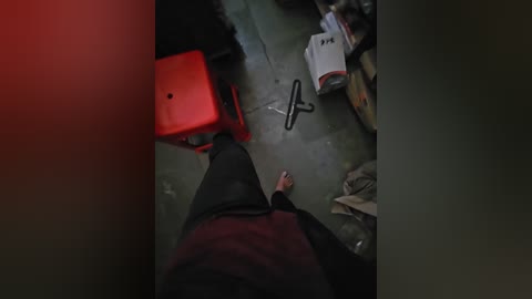 A dimly lit video shows a person's feet in black socks and dark pants standing on a concrete floor with scattered debris, including a red plastic stool, a white box, and scattered papers.