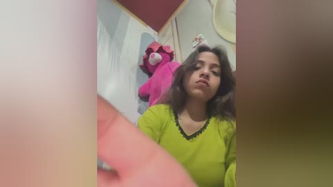 Video of a woman with shoulder-length dark hair in a lime-green sweater, standing in a dimly lit room with a large pink teddy bear in the background.