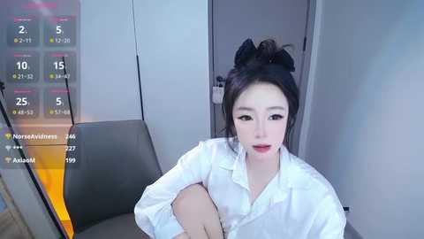 Video of an East Asian woman with fair skin, wearing a white blouse, sitting on a gray chair in a modern room. She has black hair styled in a bun.