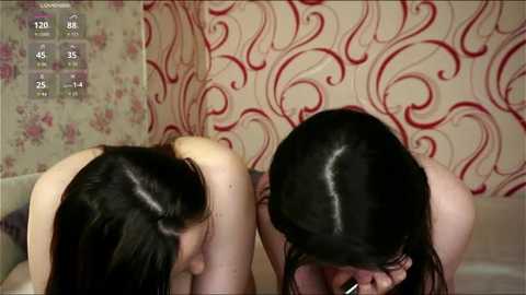 Video of two nude women with long, dark hair, kneeling on a bed with floral wallpaper, looking at each other, in a bedroom setting.