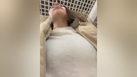 Video of a pregnant woman with light skin, wearing a beige cardigan and white top, leaning back in a laundry basket, head tilted back, eyes closed, smiling.