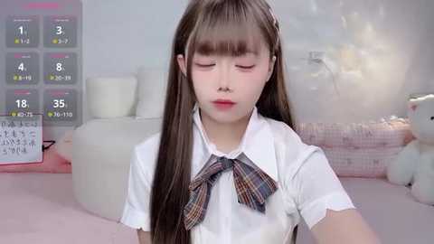 A video of a young Asian girl with long brown hair, wearing a white blouse with a plaid bow tie, sitting in a soft, pink room with plush toys and a calendar in the background.