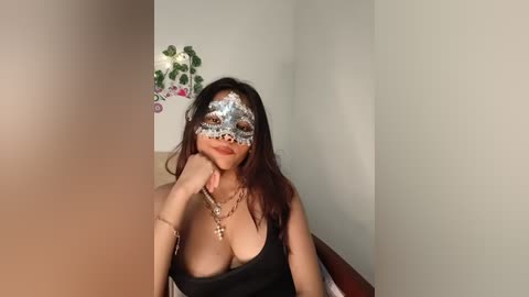 Media: Video of a woman with medium skin tone and long dark hair, wearing a silver masquerade mask, black strapless top, gold necklace, and a playful expression, in a simple indoor setting with a potted plant.