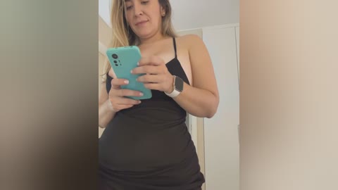 Media: A video of a plus-size woman with light skin and long blonde hair, wearing a black tank top and a smartwatch, taking a selfie in a hallway with beige walls.