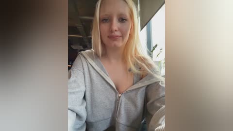 Media: Video of a young woman with fair skin, straight blonde hair, and a slight smile, wearing a grey hoodie indoors.