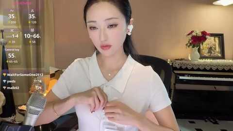 Media: Video of an Asian woman with long black hair, wearing a white blouse, sitting at a piano with a microphone. Background includes a floral arrangement, framed pictures, and a clock.