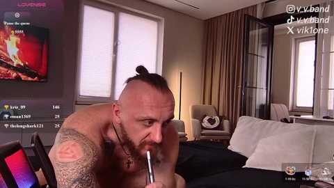 Video of a shirtless man with a topknot and tattoos, smoking, in a modern, dimly lit bedroom with a TV, bed, and windows.