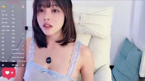 A video of a young Asian woman with straight black hair and fair skin, wearing a light blue, lace-trimmed camisole, seated in a white chair with a teal cushion. A live stream interface with chat and heart icons is visible.