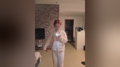 Media: A video of a woman with dyed red hair, wearing white pajamas, taking a selfie in a modern, minimalist bedroom with a circular-patterned wall, TV, and white dresser.