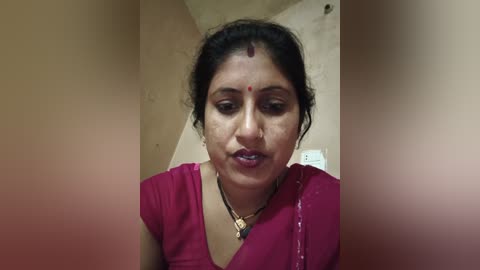 Video of a South Asian woman with medium skin tone, wearing a maroon sari, forehead bindi, and simple jewelry. She has a solemn expression. Background shows beige walls and a white electrical outlet.