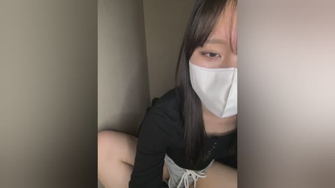 A video of a young Asian woman with long black hair, wearing a white face mask, black top, and gray shorts, crouching in a dark, confined space with beige walls.