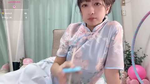 Media: Video of an Asian woman in a pastel-colored cheongsam, holding a pink balloon, with a soft-focus background of a room with light blue curtains and stuffed toys.