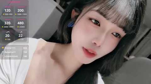 Video of a young East Asian woman with pale skin, long black hair, and large green eyes, wearing a white top, captured indoors with a smartphone screen overlay showing streaming statistics.
