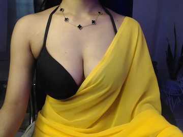 A video of a woman with fair skin, wearing a black bra and a yellow sari, adorned with a delicate necklace, sitting indoors with a dimly lit background.