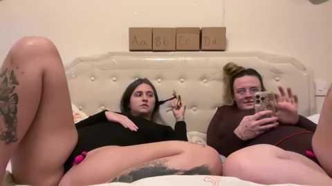 Video of two women with pale skin and tattoos, one in a black bodysuit, lying on a bed; the other in a brown sweater, holding a phone, smiling.