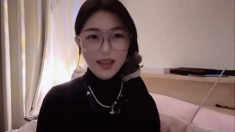 Media: Video of a young Asian woman with straight dark hair, wearing glasses and a black turtleneck, smiling in a simple, dimly lit bedroom with a white wall and a shelf holding a TV and a book.