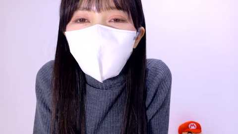 Media: Video of an East Asian woman with straight black hair and bangs, wearing a gray ribbed turtleneck, white surgical mask, and standing against a plain white background.
