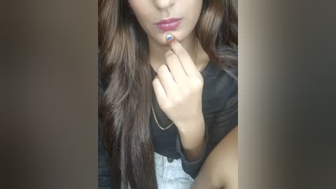 Media: Video of a young woman with long, wavy brown hair, light skin, wearing a black top and light blue jeans, biting her fingernail, with a blurred background.