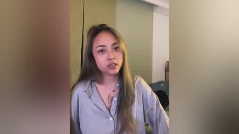 Video of a young woman with long, wavy brown hair, wearing a light gray button-up shirt, standing in a dimly lit room with beige and green walls.
