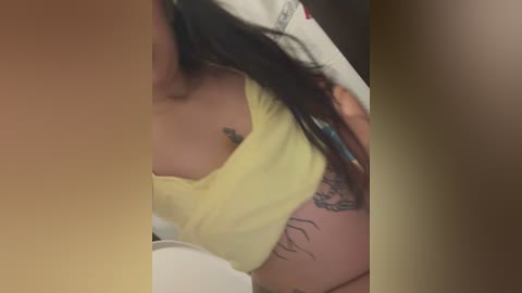 Video of a partially obscured woman with light brown skin, wearing a yellow tank top, showing a tattoo on her left arm, taken in a bathroom.