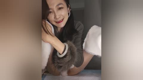 Video of a smiling Asian woman with long black hair, wearing a gray fur-trimmed sweater, white gloves, and beige pants, crouched in a dimly lit room, peering through a slit in a white curtain.