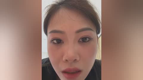 Video of an East Asian woman with fair skin, brown eyes, and straight brown hair, wearing subtle makeup, set against a blurred, beige background.