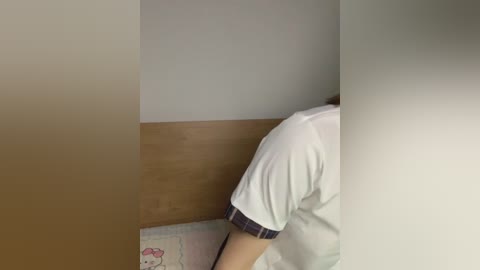 A video of a person's upper back and shoulders, wearing a white T-shirt with a plaid trim, standing in a room with beige walls and a pink Hello Kitty cushion.