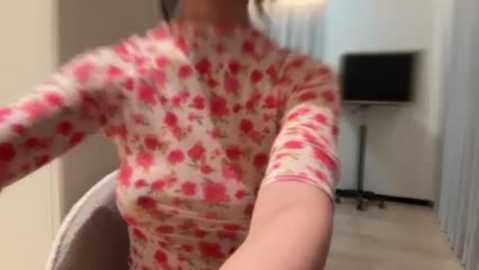 A video of a woman wearing a tight, floral-patterned, long-sleeve top in a narrow hallway with a flat-screen TV on the right.