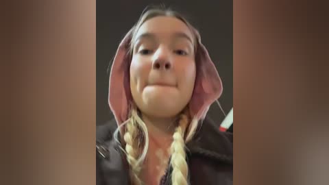 A close-up video of a young girl with light skin and long blonde hair, styled in two braids, wearing a black jacket. She has a slight smile, with a pink hooded sweatshirt partially covering her head. The background is dark and out of focus.