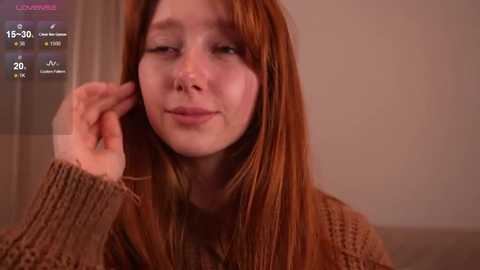 Video of a young woman with fair skin and long, straight auburn hair, wearing a textured brown sweater. She looks relaxed, with one hand resting near her face. Background is a muted beige.