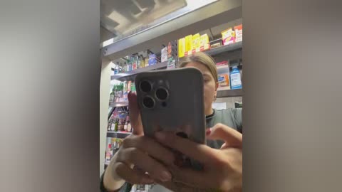 A video of a woman with light skin and blonde hair taking a selfie in a well-lit grocery store. She is holding a gray iPhone with three cameras, partially obscuring her face. The background reveals colorful shelves stocked with various products.