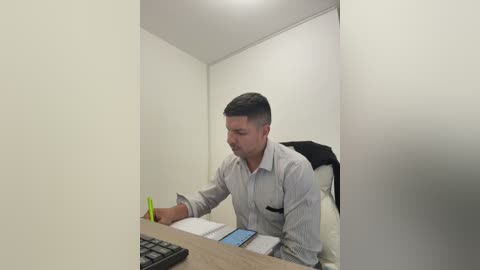 Video of a man with short dark hair, wearing a light blue striped shirt, typing on a laptop at a desk in a small, dimly lit room with white walls.