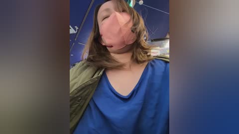 Media: Video of a person wearing a pink surgical mask, blue scrubs, and an olive green jacket, with their eyes closed and mouth open, in a medical setting with blue walls and hanging equipment.