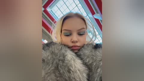 Video of a young woman with light skin and blonde hair, wearing dramatic green eyeshadow and a fur coat, indoors with a geometric red and white ceiling.
