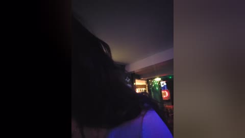 A dimly lit video captures a person's dark hair and shoulder in the foreground, with neon lights and a green screen in the background, creating a moody, slightly blurry atmosphere.